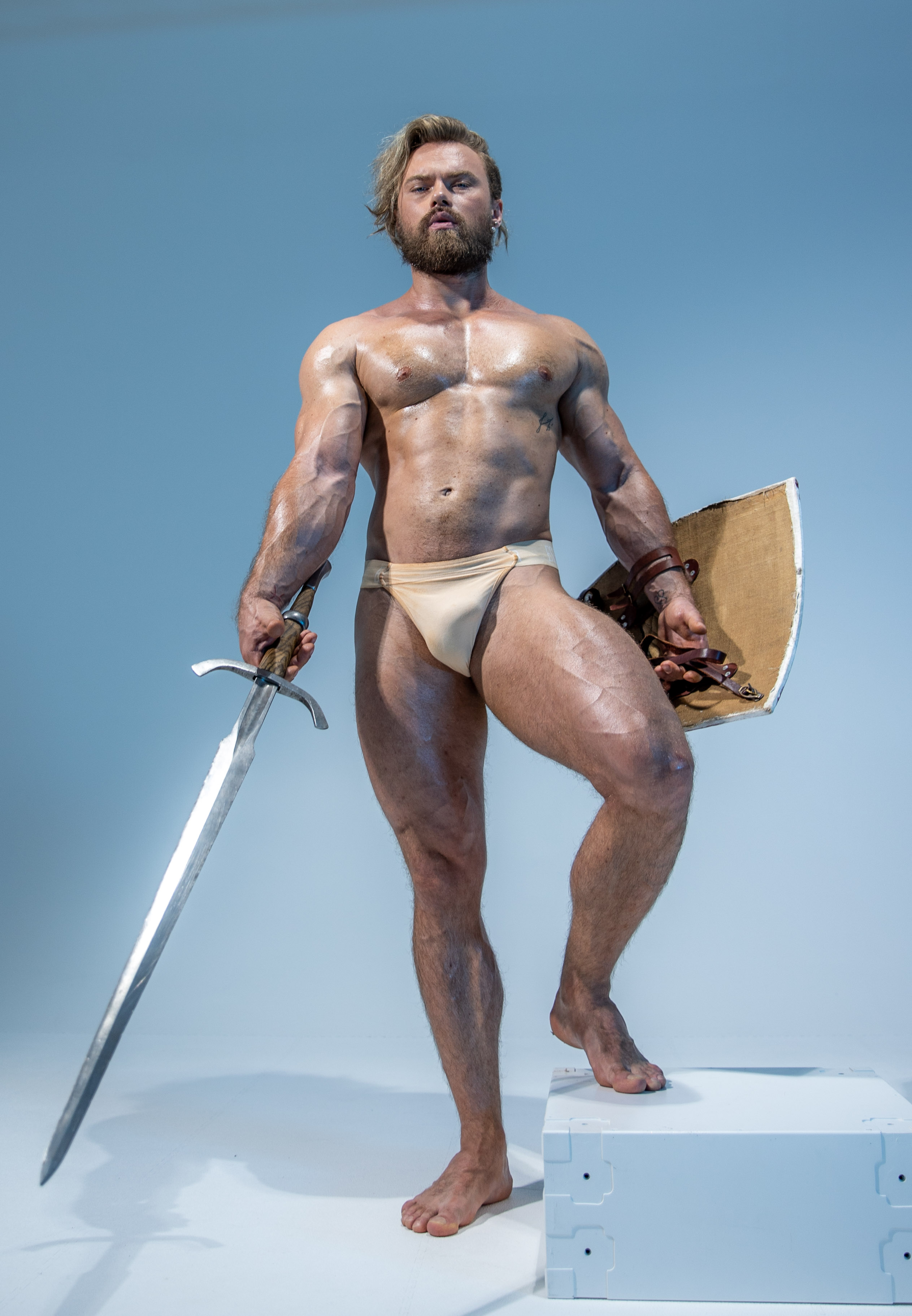 a muscular man with long blond hair stands on a white pedestal in a confident pose. he is wearing a loincloth and holds a large sword in his right hand and a round shield in his left. he is illuminated by soft studio lighting against a light blue background. the scene evokes a sense of power and strength, reminiscent of a fantasy warrior or historical figure.