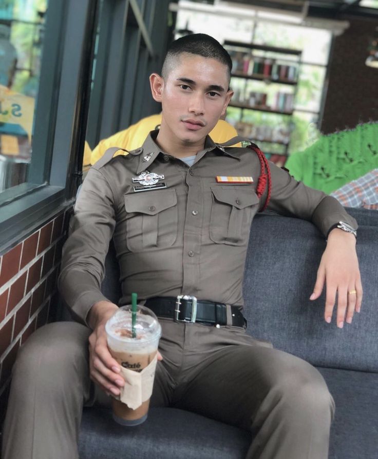 a handsome asian police officer in uniform is sitting on a couch and relaxing. he is holding an iced coffee and looking at the camera. the background is blurred out.
