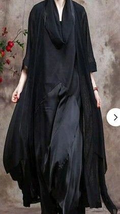 a woman in a black, longsleeved dress stands in front of a gray wall. she is posing for a fashion photograph. the dress is loosefitting and has a cowl neck. it is stylish and elegant.  the woman is wearing her hair down. she is looking at the camera with a serious expression on her face. the background is out of focus.  there is a plant on the left side of the frame.  the image is welllit and the woman's features are clear.