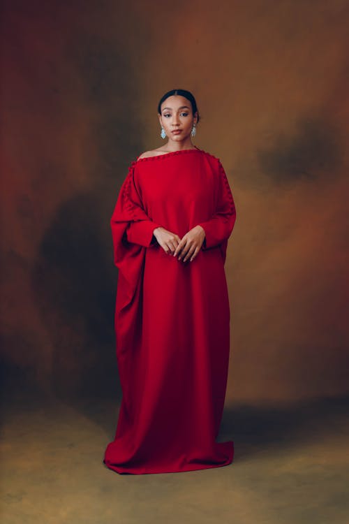 a fulllength studio portrait shows a woman wearing a long red gown with long sleeves and a oneshoulder design. she has her hands clasped gently in front of her. she is wearing large turquoise earrings. the backdrop is a mottled brown and olive green.
