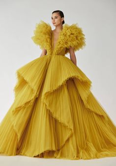 a woman in a stunning yellow tulle gown with ruffled detailing. the gown features multiple layers of pleated tulle and has a plunging neckline. the model is posing with her hair pulled back, showcasing the intricate details of the dress. the dress has a formal and elegant look, perfect for a special occasion. the yellow color is vibrant and eyecatching, making the dress a true statement piece.