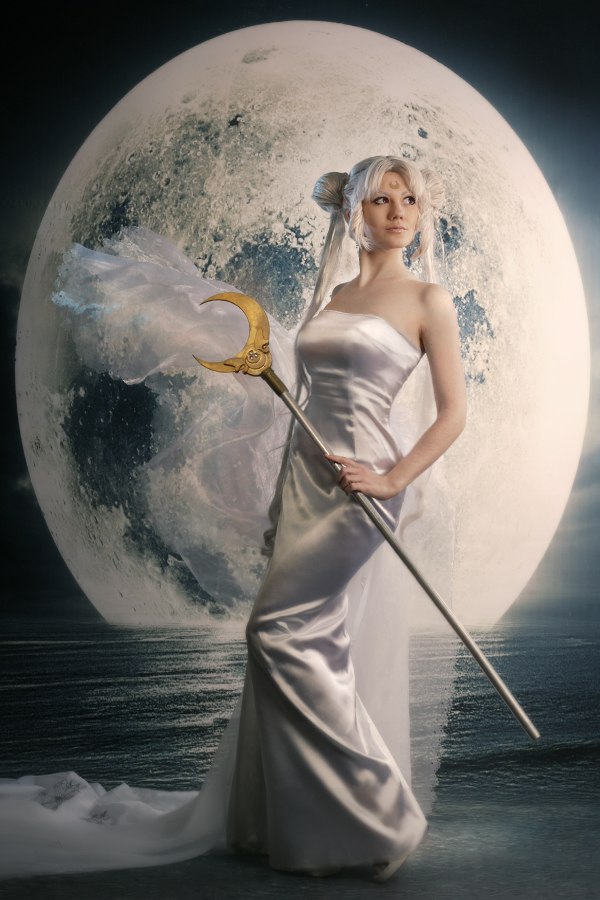 a cosplayer dressed as princess serenity from sailor moon stands in front of a large moon. she is wearing a long, silver dress and a tiara, and she is holding a staff with a golden crescent moon on top. she has fair skin and blonde hair that is styled in a bun. the background is a dark blue color with a body of water.