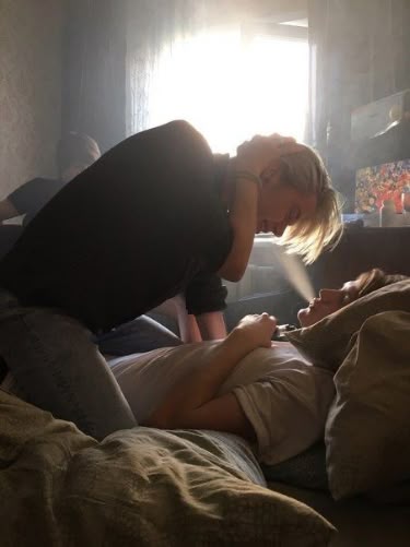 a photo shows a couple indoors. one person is lying down on a bed or couch with pillows, while the other person is kneeling or standing over them, hugging them. there is smoke or vapor coming from the mouth of the person lying down. the room appears to be lit by daylight coming through a window. a third person is in the background.