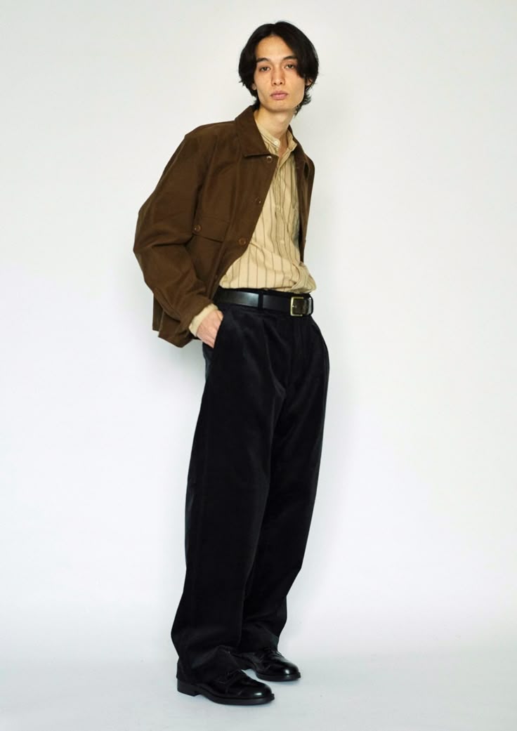 a full shot of a man standing in a studio wearing a brown corduroy jacket over a tan striped shirt, black corduroy trousers, and black dress shoes with a black belt. he has his hand in his pocket and is looking at the camera.