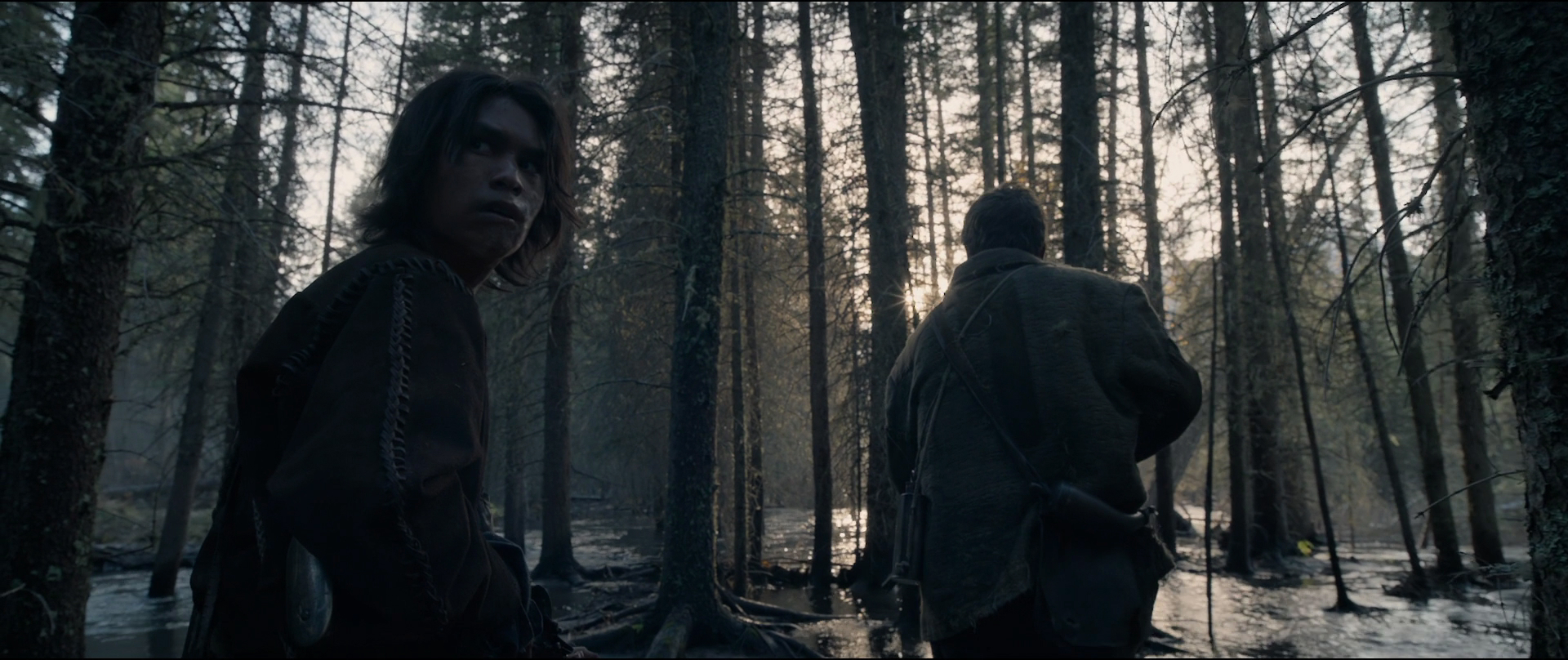 two men are standing in a dark forest. one man is looking towards the left of the frame while the other is looking straight ahead. the forest is thick with tall trees and the sunlight is filtered through the leaves, creating a shadowy and mysterious atmosphere.