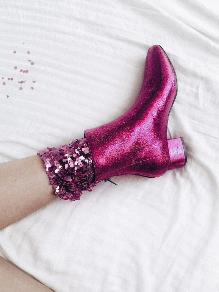 a closeup shot of a woman's leg wearing a pink metallic ankle boot with a block heel. the boot is paired with sparkly pink sequined socks.  the scene is set against a white, textured background.  the overall style is glamorous and festive.