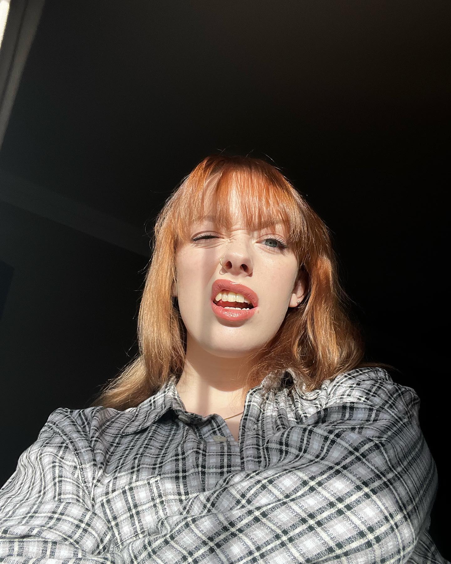 loseup selfie of a young woman with bright red hair styled in bangs. she is wearing a grey and white plaid shirt and has a playful facial expression, perhaps winking, with her mouth slightly open. sunlight highlights one side of her face, creating a contrast with the darker background.  she has a nose piercing visible. the overall mood is cheerful and casual.