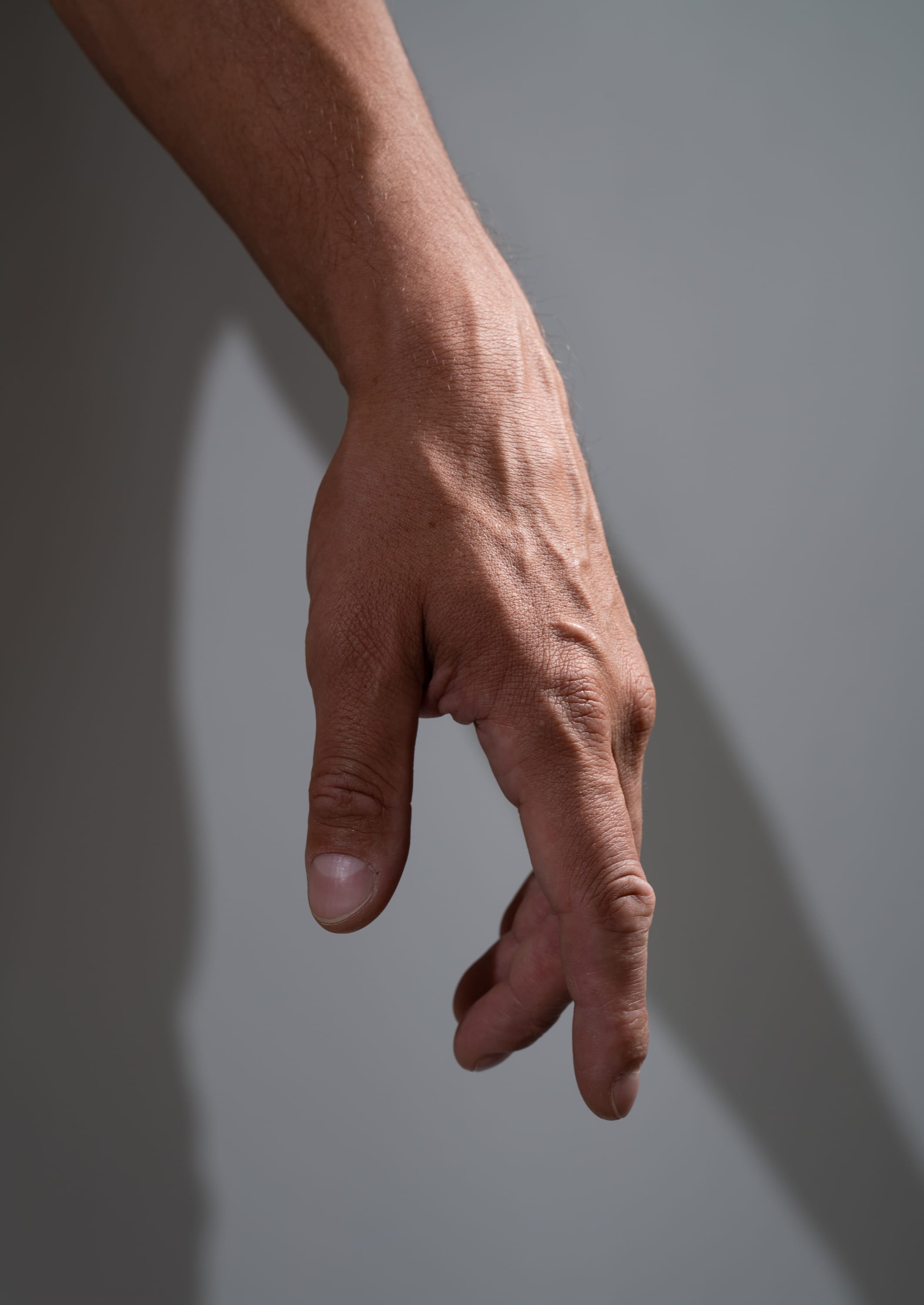 a closeup shot of a man's hand and arm extended, lit by hard, side lighting which creates a distinct shadow on the background. the hand is relaxed with a slight curl to the fingers. the image highlights the details of the skin texture and veins.