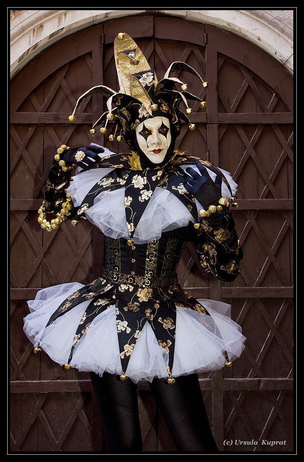 a person in a black and gold jester costume with a white mask, standing in front of a wooden door with diamondshaped panels. the costume includes a jester hat with bells, a ruffled collar, a bodice, a tulle skirt, and gloves with golden adornments. the mask has black diamond shapes around the eyes and red lipstick.