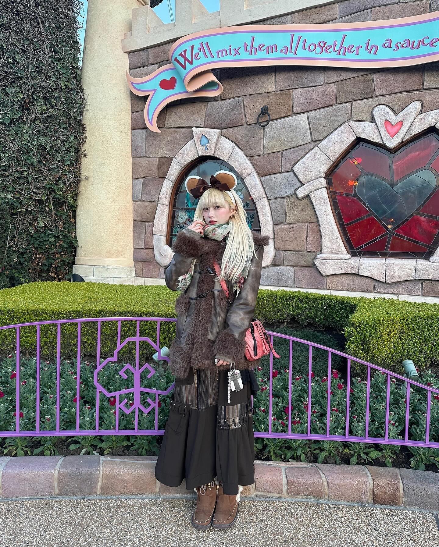 the image shows a young woman standing in front of a whimsical alice in wonderland themed wall at a disney park, possibly tokyo disneyland. she's wearing a brown faux fur coat, a long darkcolored layered skirt, brown boots, and a minnie mouse ear headband.  she carries a small reddishbrown handbag. the wall behind her features a quote, "we'll mix them all together in a sauce," and has stained glass windows with heart shapes. the scene is framed by a purple metal fence and lush greenery.  the overall aesthetic is autumnal, and the woman's style is  cute and fashionable, possibly influenced by japanese street style.