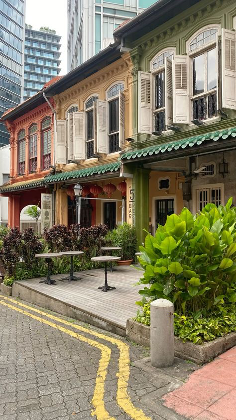 the image shows a row of colorful shophouses in singapore. the shophouses are traditional asian architecture with distinctive features like decorative facades, windows with shutters, and green roofs.  the buildings are situated along a street with a paved area and yellow lines. the foreground includes outdoor seating at a cafe or restaurant, along with lush green plants and a concrete bollard.  in the background, modern skyscrapers are visible. the overall scene contrasts old and new architecture in a vibrant urban setting.