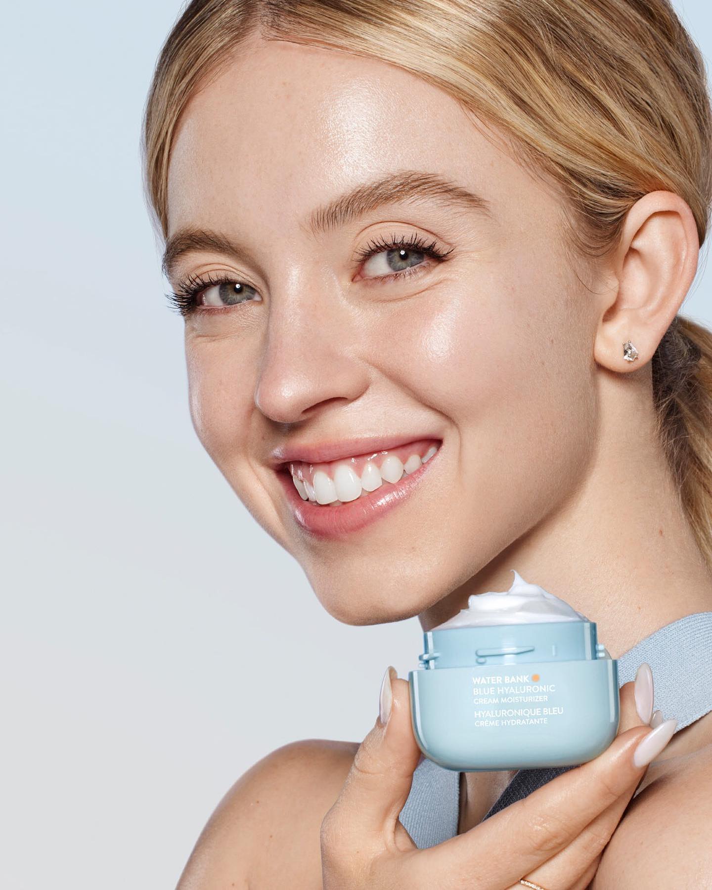 a closeup shot features a smiling young blonde woman holding a jar of laneige water bank blue hyaluronic cream moisturizer. the woman's skin appears radiant and healthy. the focus is on both the woman's happy expression and the product she is holding, highlighting its association with beautiful and hydrated skin.  the background is a soft, light blue, keeping the focus on the model and the product.