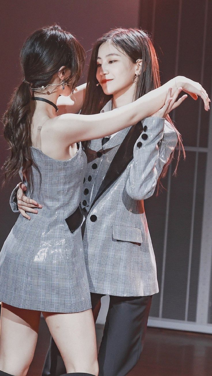 two young women are dancing together on a stage. the woman in the front is wearing a plaid dress and the woman in the back is wearing a suit jacket. they are both smiling and seem to be having a lot of fun. they are likely friends or a couple.