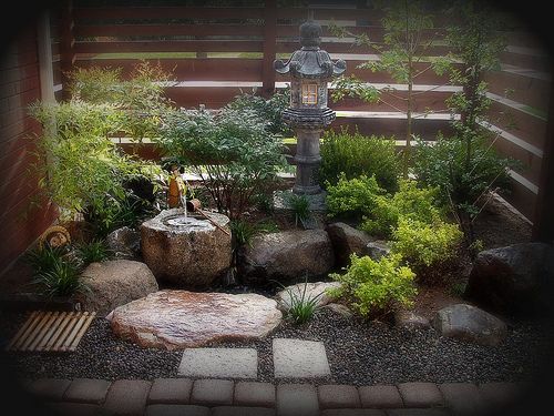 the image showcases a wellmaintained zen garden, featuring a stone lantern as a focal point. a water fountain and various rocks and pebbles are strategically placed amidst lush green plants and shrubbery. stepping stones guide visitors through the garden, which is enclosed by a fence, creating a peaceful and tranquil outdoor space.