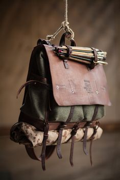 a vintagestyle backpack made of leather and canvas is suspended from a rope against a blurred background. the bag features multiple straps, a roll at the bottom, and a flap closure. the aesthetic suggests outdoor adventure and travel.