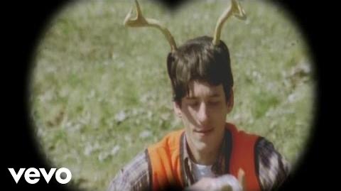 a young man wearing a pair of antlers looks down and holds something out of frame, while a camera with a blurry background, presumably showing a forest or woods, shows a small portion of the man's face and a vevo logo at the bottom. the man is outdoors and seems to be participating in a hunting activity.