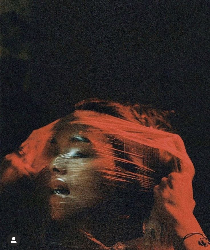 the image is a closeup, moody portrait of a person's face partially obscured by a sheet of translucent plastic wrap. the lighting is predominantly red, casting dramatic shadows and creating a dark, mysterious atmosphere. the plastic clings to the face, obscuring details while highlighting the person's features. the hands of another person are visible, manipulating the plastic around the face. the overall aesthetic is artistic and sensual, suggesting themes of mystery, constraint, or vulnerability.  the photograph's style suggests a cinematic or fashion photography approach, with a gritty and alternative tone.