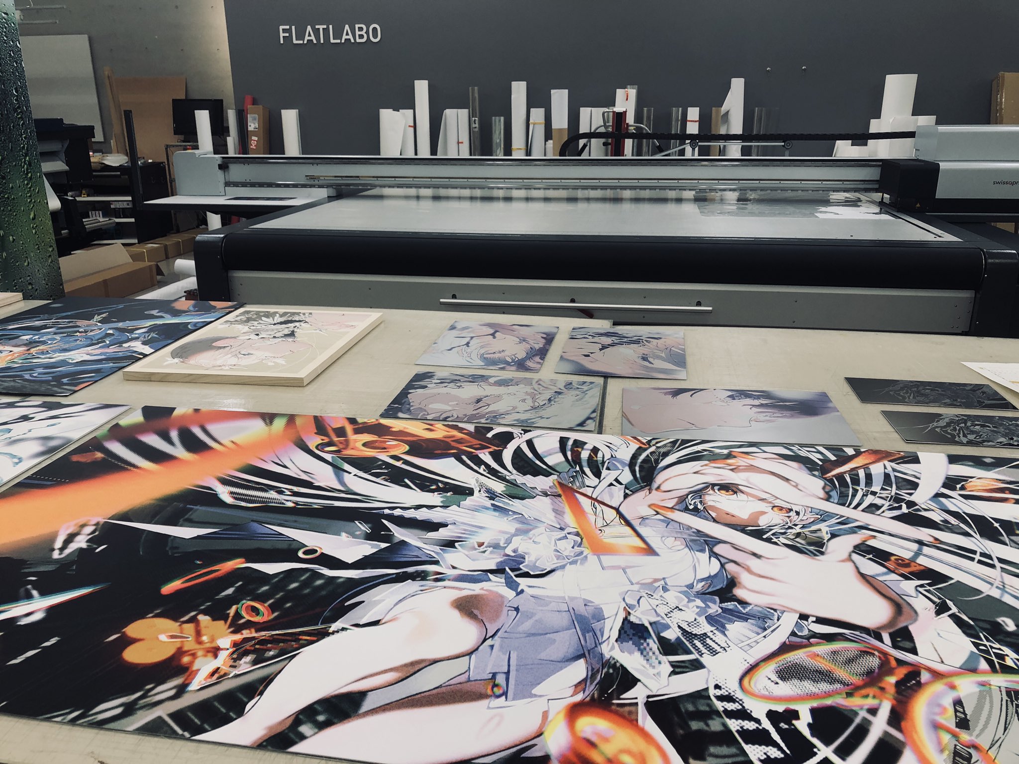 a large format printer sits on a table in a print shop.  several printed posters are laid out on the table in front of the printer.  the posters feature colorful animestyle graphics.