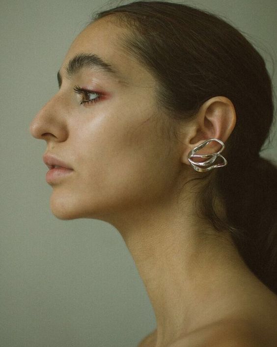 a closeup profile shot of a woman with dark hair pulled back into a low ponytail. she's wearing large, silver sculptural earrings. her skin is smooth with subtle makeup highlighting her eyes and lips. the background is a muted greenishgray.