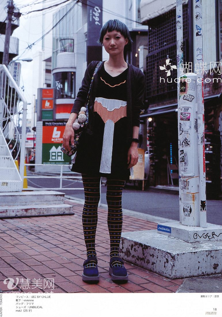 a young woman with short blue hair is standing on a city sidewalk, posing for a street style photo. she's wearing a black dress with an orange and creamcolored panel in the front. she is also wearing black and yellow patterned tights and black platform shoes. she has a black purse with a white pompom hanging from her right shoulder. behind her is a city street scene with buildings and signs in japanese. the photo is welllit, as if taken on a sunny day.