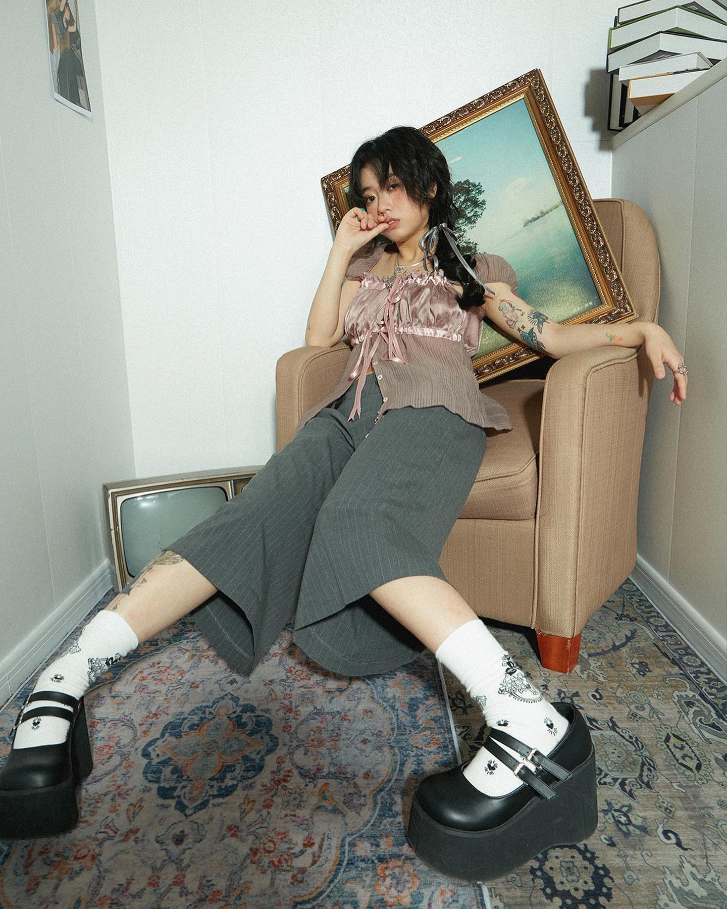a young asian woman with dark hair sits in a vintage armchair. she's wearing a pink crop top, grey wideleg pants, white ankle socks and black platform mary jane shoes.  an antique picture frame is leaning against the chair. the overall aesthetic is vintage and stylish.
