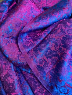 closeup shot of a luxurious purple and blue brocade fabric with a floral pattern. the fabric is draped and folded, creating a beautiful display of its texture and shine. the image captures the intricate details of the pattern, showcasing the craftsmanship of the fabric. the rich colors and elegant sheen make it a perfect choice for formal occasions such as weddings or parties.