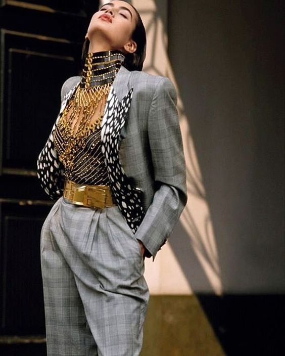 a fashion model poses for a photograph. she is wearing a gray blazer, gray pants, and a gold belt. she has on a large gold necklace with a choker style top. she is looking up with her eyes closed. the background is a plain gray and white wall.