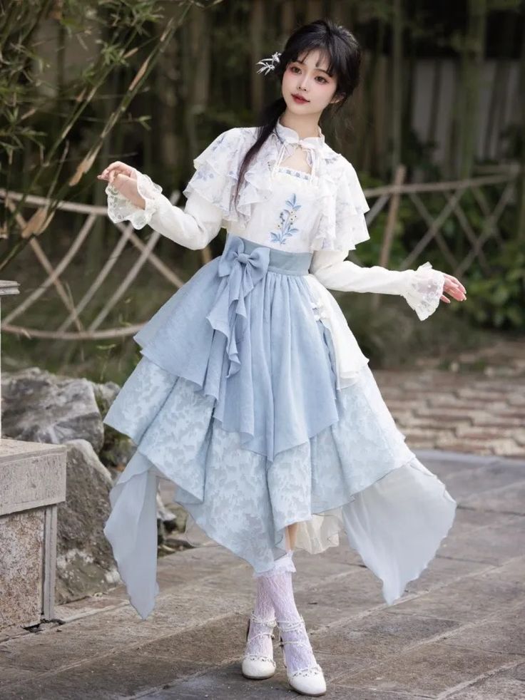 a young woman poses gracefully in a garden setting. she is wearing a blue and white lolita fashion dress. her outfit features a white blouse with lace details and a blue skirt with multiple layers and a floral pattern. a white shawl drapes over her shoulders. the overall look is elegant and feminine. the environment features lush greenery and a stone fence. the image is welllit with natural daylight.