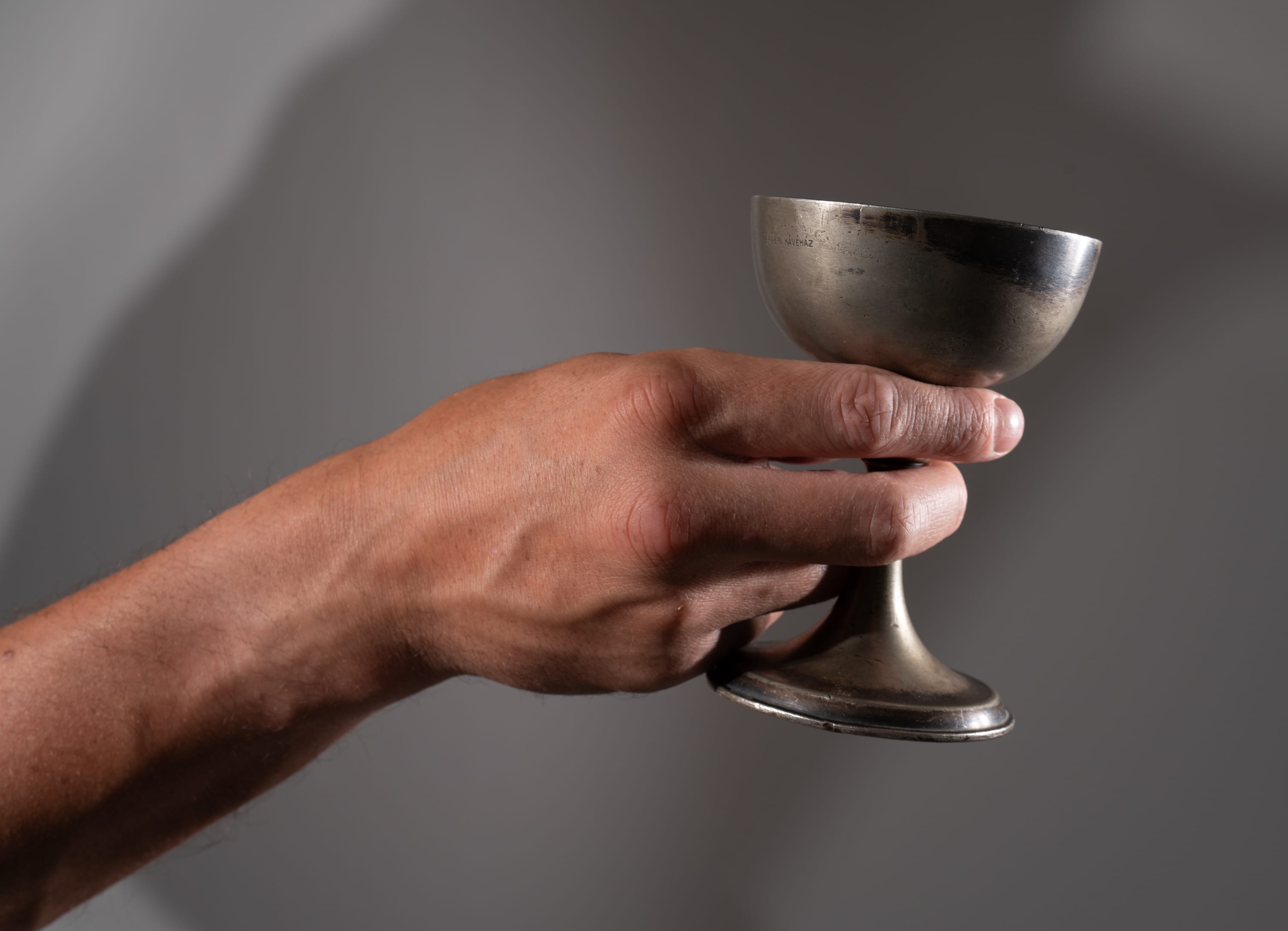 a man's hand holds a tarnished, silvercolored cup against a gray background.