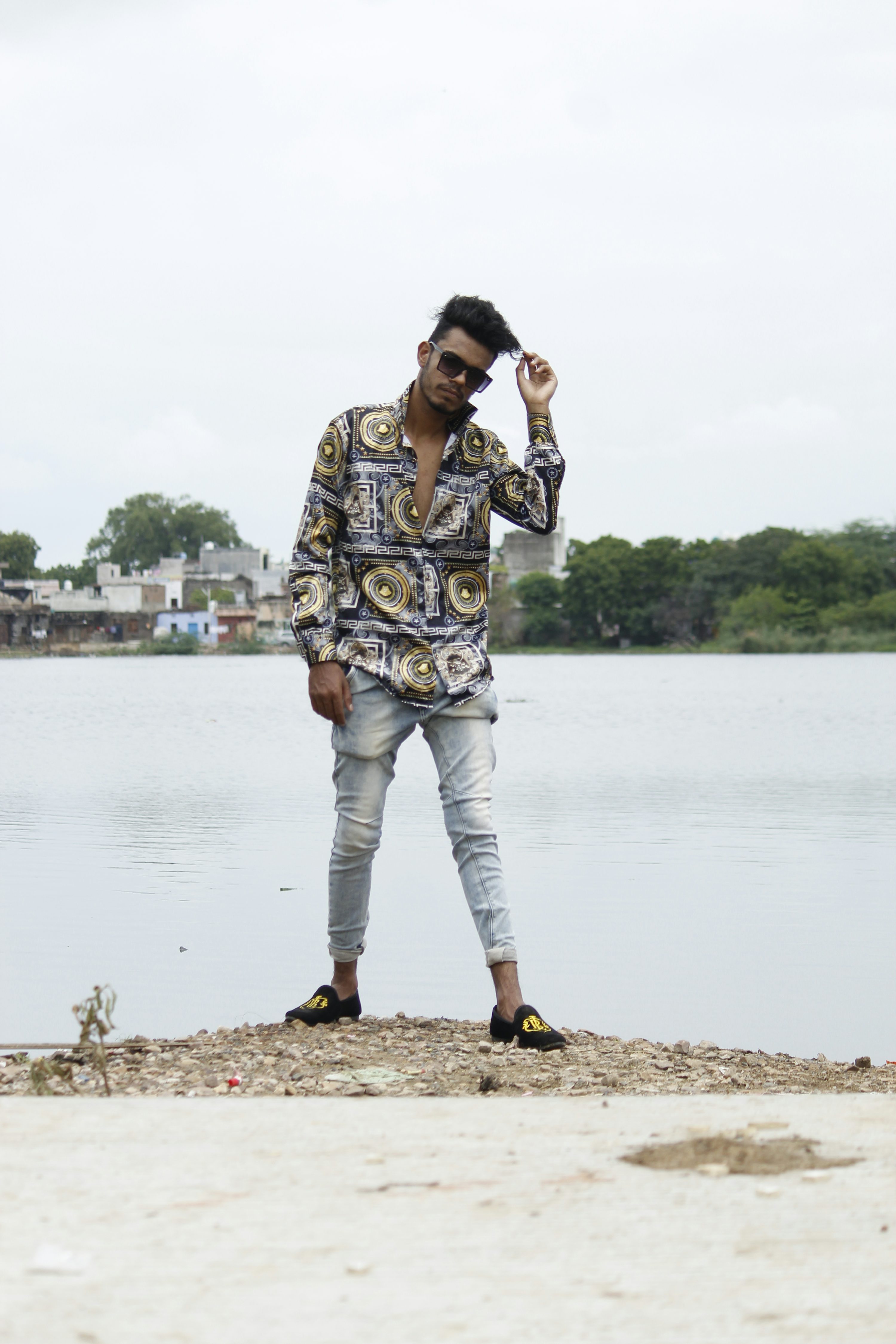 a young man stands confidently by a calm lake, posing for a photo. he is wearing a stylish patterned shirt, lightwash jeans, and black loafers.  he has dark hair and is wearing sunglasses, which he is adjusting with one hand. the background features a calm body of water and a distant shoreline with trees and buildings. the overall mood is casual and relaxed.