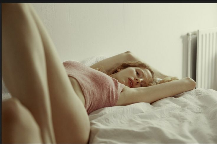 a woman with auburn hair is lying on a bed with a white sheet. she is wearing a pink shirt and has one arm raised above her head. a white radiator is visible in the background.