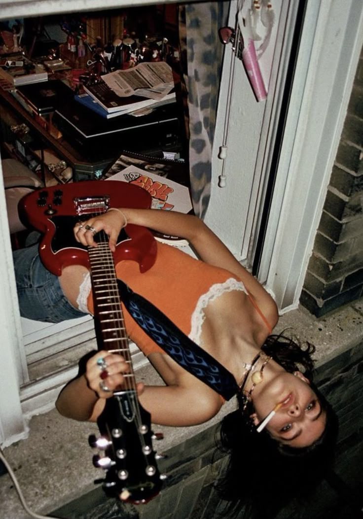a woman is lying on a windowsill, holding a guitar and smoking a cigarette. she is wearing an orange tank top with white lace trim, blue jeans, and multiple rings and necklaces. her hair is dark and curly. the windowsill is part of an open window with a view into a messy room filled with books, papers, and other items. the guitar is red with a black headstock, and it has a blue strap with a flame design. the window is white and appears to be part of a brick building.