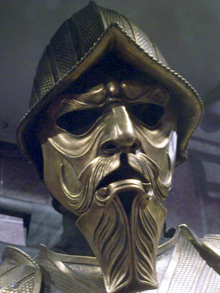 the image shows a golden helmet with a face mask and beard, part of a medieval armor set. the helmet is shaped like a conical helmet with a face mask and a stylized beard. the armor has a golden color. it could be part of a costume or cosplay outfit.