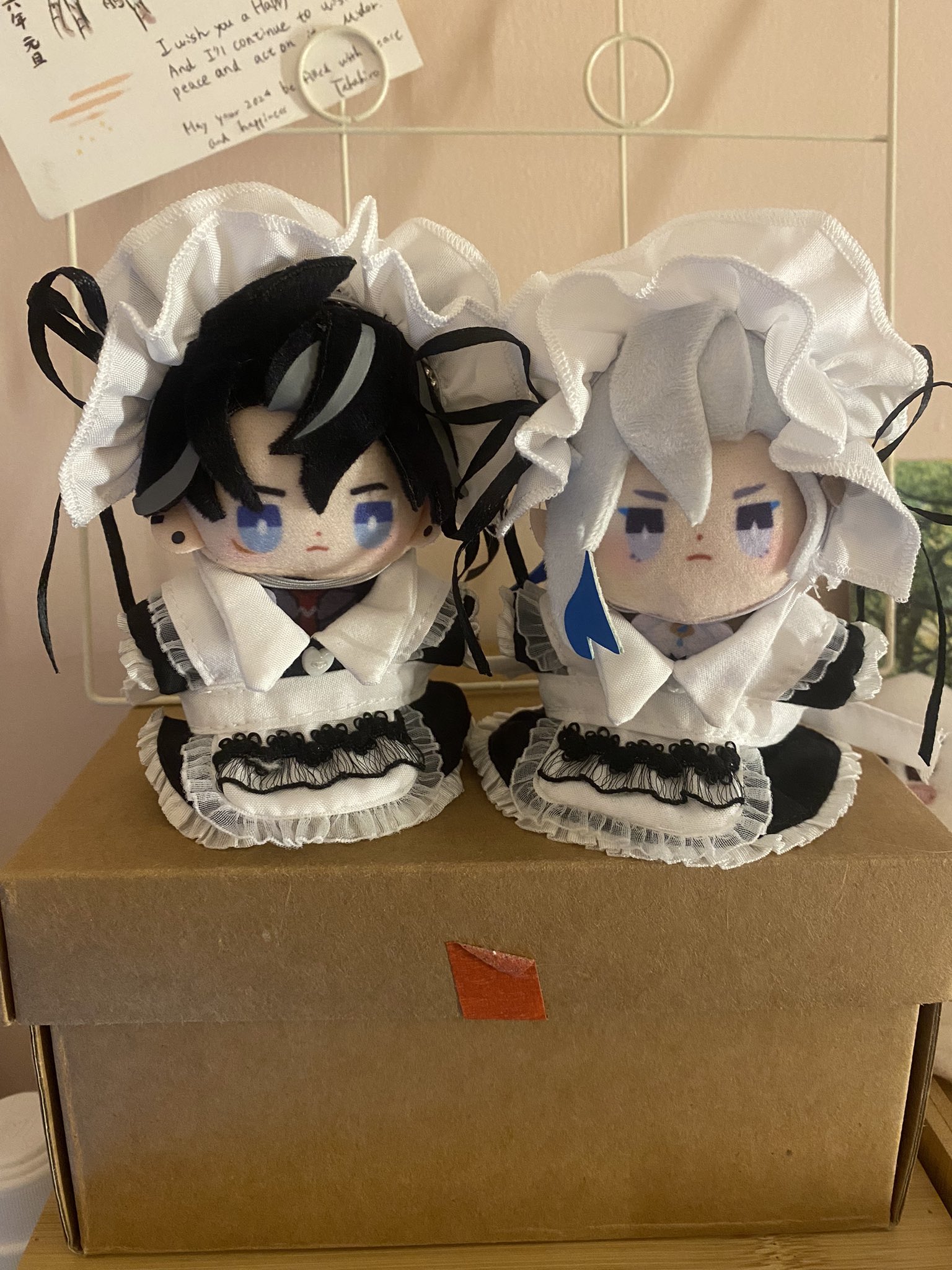 two anime plush dolls dressed in maid outfits, one with black hair and one with white hair. they are both standing on a brown box. the black haired doll is on the left and the white haired doll is on the right. both dolls have the same outfit, which consists of a white dress with black trim, a black apron with a white lace trim, and a white headband with black trim. the dolls have the same face, which is very cute and kawaii. the dolls are both looking forward.
