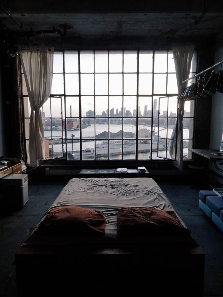the image shows a minimalist bedroom in a loft apartment with a large window offering a stunning view of the manhattan skyline. the room is sparsely furnished, featuring a bed with brown pillows, and a mostly empty floor. the large window is divided into many panes and has white curtains partially drawn. the overall atmosphere is calm and serene, highlighted by natural light streaming in from the window. the industrial aesthetic of the loft is evident in the exposed pipes, concrete ceiling, and general design.  the city view is the focal point, dominating the background of the image.