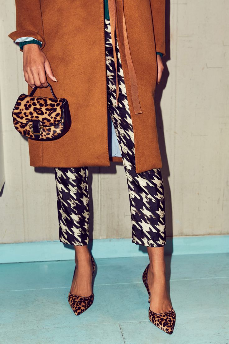 a stylish woman is shown from the knees down. she is wearing a camelcolored coat, houndstooth trousers, and leopard print shoes and handbag.  she is standing on a light blue floor against a light beige wall.  the overall style is autumnal/winter.