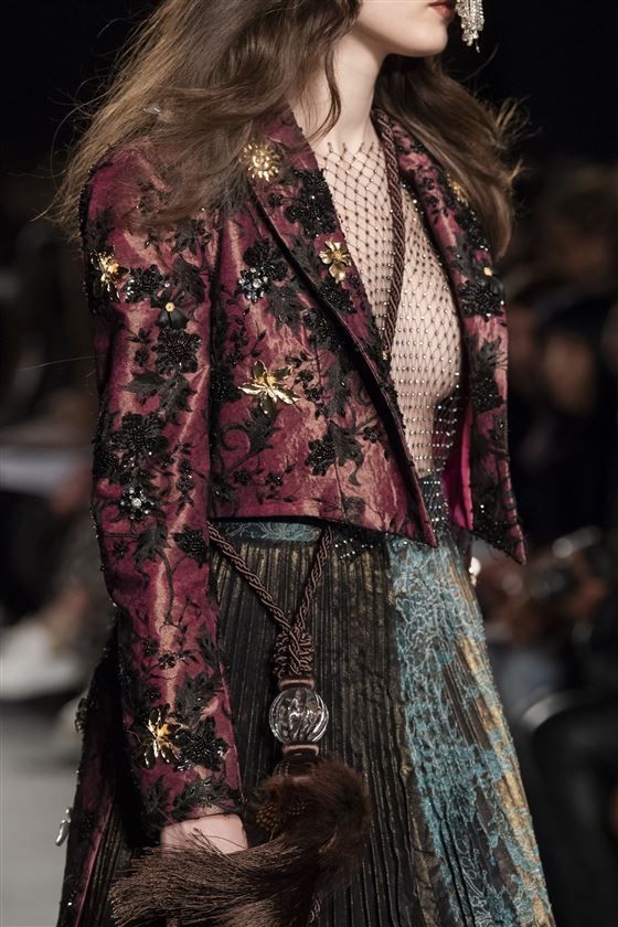 loseup view of a female model on a fashion runway wearing a richly detailed dark red embroidered jacket and a pleated skirt. the jacket is adorned with intricate beading, sequins, and gold accents.  the skirt is a mix of dark colors and textures. the model has long brown hair and is wearing a sheer, delicate top underneath the jacket. she carries a dark fur accessory attached to a cord and has ornate earrings. the overall aesthetic is glamorous and sophisticated. the image showcases the high level of detail and artistry in the clothing design.
