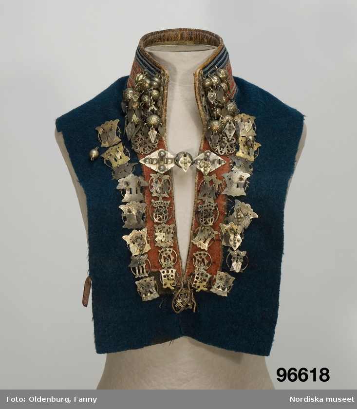 a closeup studio shot features a mannequin dressed in a traditional folk costume vest. the vest is primarily dark blue with a contrasting orange or red panel running down the front. the collar is adorned with a striped pattern of blues, oranges, and whites. decorative metalwork, including silvercolored pendants and embellishments, is heavily featured on the vest, adding an ornate and detailed appearance to the garment. the item is housed at nordiska museet.