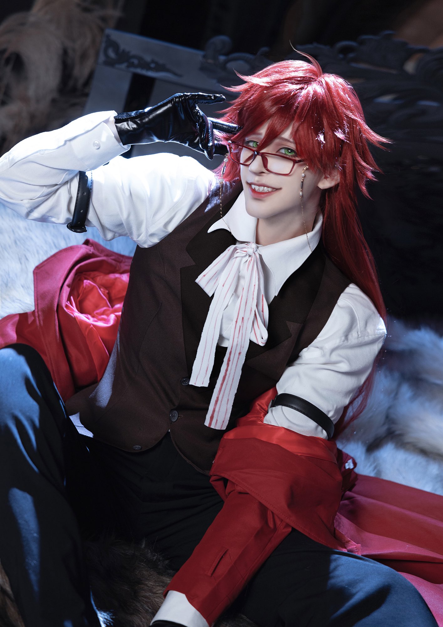 a person is cosplaying sebastian michaelis from black butler. they are wearing a black vest, white shirt, and red coat. they have red hair, black eyes, and are wearing glasses. they are looking at the camera and sitting down. the background is blurry and out of focus.