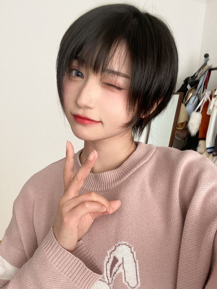 a young asian woman with short black bob hair winks at the camera with a sweet smile. she is wearing a pink sweater and her hair is styled in a cute bob cut.