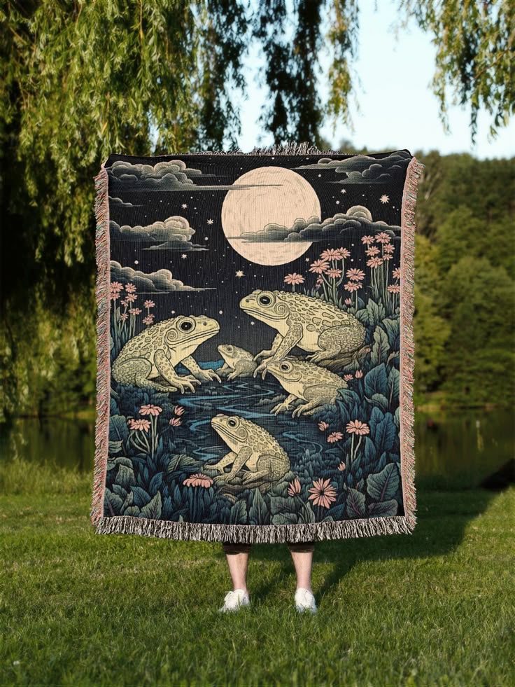 a person stands on a grassy lawn in an outdoor setting, holding up a woven blanket with a naturethemed design. the blanket features a nocturnal scene with a full moon, stars, clouds, and illustrations of frogs or toads in a pond or marshlike environment with flowers. the person's legs and shoes are visible behind the blanket. the background includes trees, foliage, and a body of water, suggesting a park or natural landscape. the blanket is held up high enough that only the legs and shoes are visible of the person.