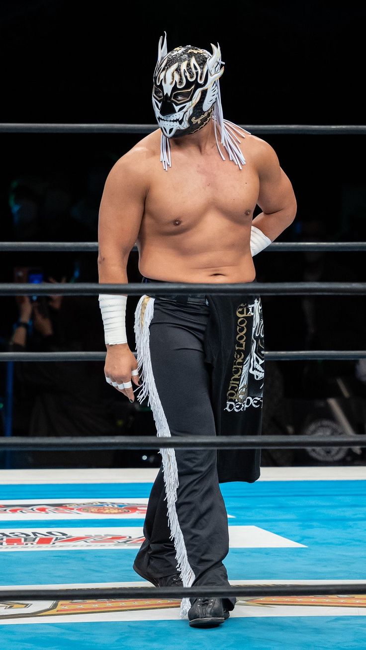 a masked professional wrestler, possibly lucha libre style, stands inside a wrestling ring. the wrestler is shirtless and wears black wrestling pants with white tassels and gold detailing. the mask is predominantly black and white with a dragonlike design. the wrestler's physique is muscular and athletic. the background is blurred but shows the ropes of the wrestling ring and a glimpse of spectators in the arena. the overall ambiance suggests a wrestling event is in progress.