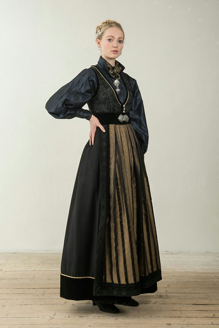 a fulllength studio shot of a woman wearing a traditional norwegian bunad. the bunad is predominantly black and gold, featuring a long skirt, a fitted vest, and a dark blouse. the woman has her hair styled in an updo and is wearing jewelry that complements the bunad.  she is standing on a wooden floor against a lightcolored background. the image portrays the elegance and detail of the traditional norwegian attire.