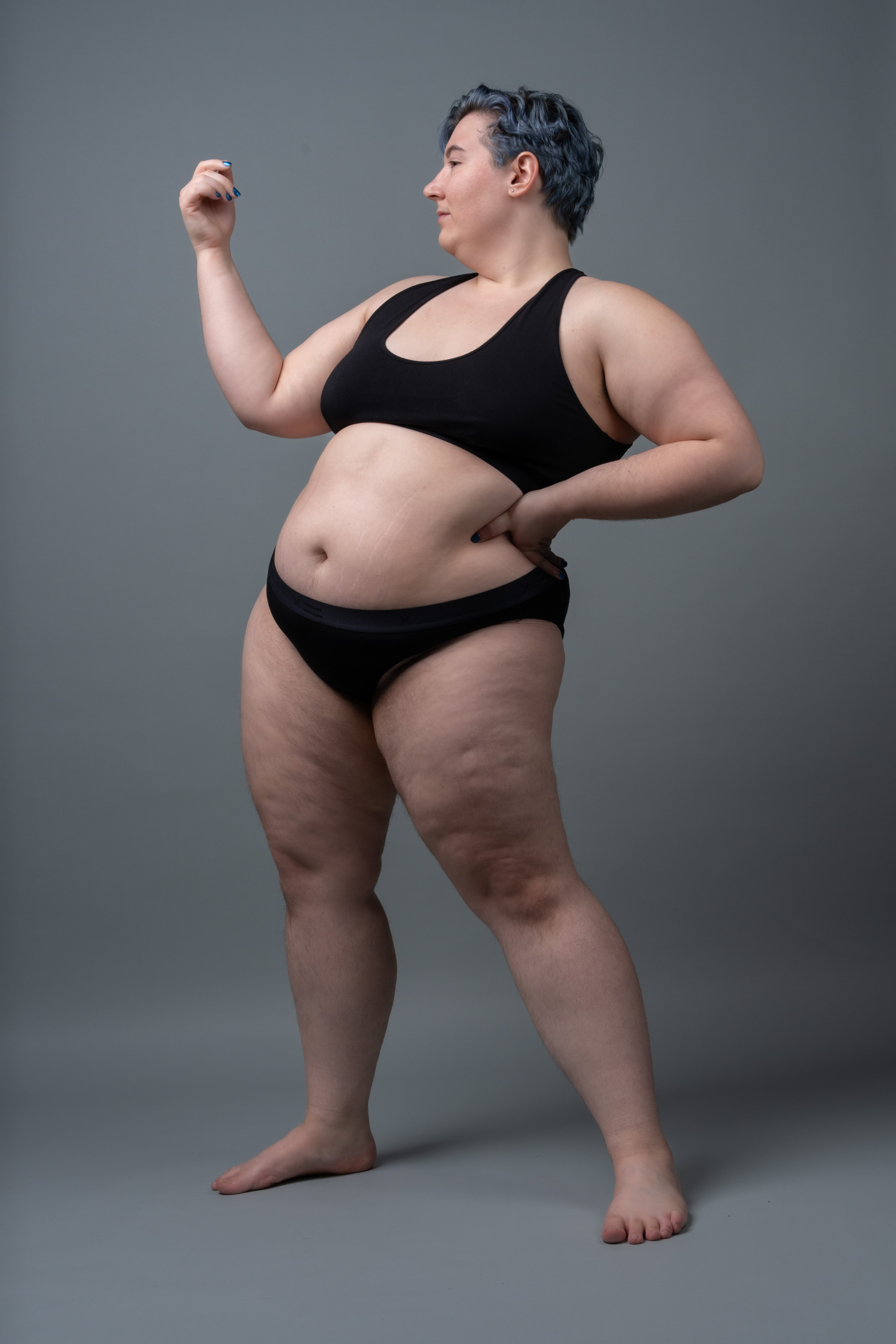 a plussize woman stands barefoot in a studio against a grey background. she is wearing black lingerie (bra and panties). her hair is styled in a short blue cut. she is looking to her left and holding a small object in her raised right hand. she has one hand on her hip and is in a confident pose. she is a fullbodied woman with cellulite visible on her thighs. the shot is a midlength, fullbody view. the overall theme is body positivity.