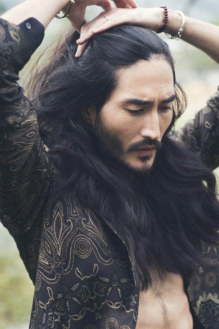 an asian man with long black hair and a beard is shown from the chest up, looking down with his hands raised and touching his hair. he is wearing a patterned black and gold jacket that is open in the front. he is also wearing bracelets and rings. the background is blurry and appears to be outdoors.