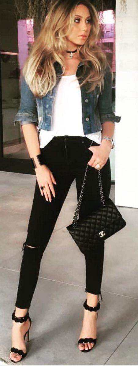 a fulllength shot of a stylish woman standing confidently. she's wearing a light blue denim jacket over a white tshirt, paired with black ripped skinny jeans and elegant black highheeled sandals. she is carrying a black quilted chanel handbag with a silver chain and logo. accessorized with a black choker necklace, a black watch or bracelet, and rings. she has long blonde hair and a welldefined makeup look. the setting appears to be an outdoor urban area with neutral tones and blurred elements in the background.