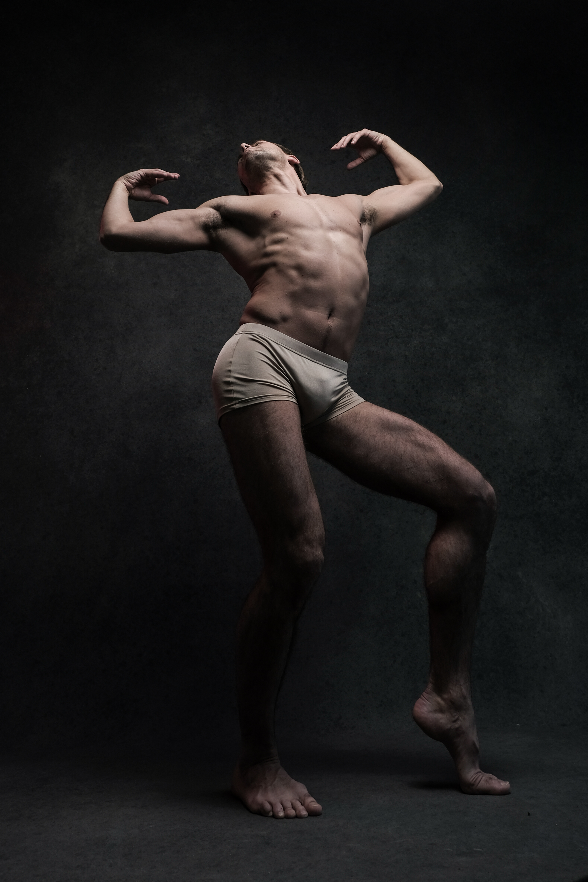 a male dancer in underwear strikes a pose against a black background in a studio. the lighting highlights his muscular physique and the graceful lines of his body.