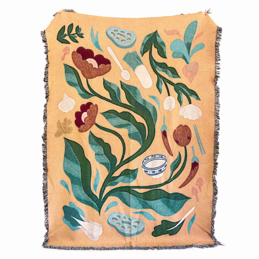 a woven blanket featuring a botanical and vegetable illustration in a colorful, decorative style. the pattern includes flowers, leaves, garlic, peppers, a bowl, a spoon, various asian vegetables, and crosssections of lotus root. the blanket has a fringed edge.
