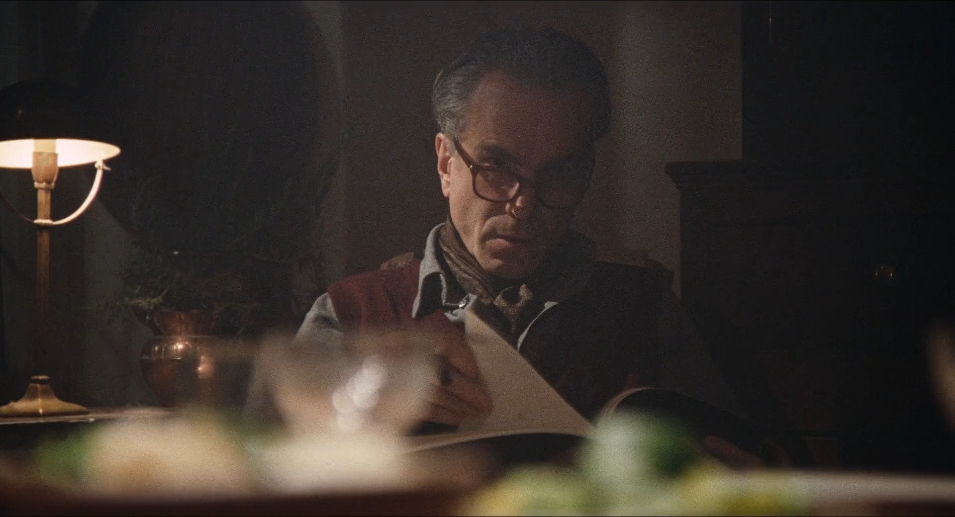 a man is sitting at a table in a dimly lit room, reading a book. the man is wearing glasses and has a serious expression on his face. a lamp is lit behind him, casting a warm glow on his face and the book. the table is made of wood and is covered with a white tablecloth. there are plants on the table, out of focus, behind the man. the room is dark, with only the lamp and the light coming from the window providing illumination. the man's glasses reflect the light from the lamp, adding to the feeling of mystery and intrigue. the man's focus on the book suggests he is completely engrossed in its contents.