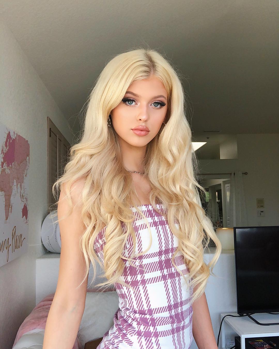 a portrait of a young woman with long blonde wavy hair, blue eyes, and wearing makeup. she is wearing a white and purple plaid dress. she is standing in a room with a map on the wall and a tv in the background. she is also wearing a necklace and earrings. the woman is loren gray. there is also a couch in the image.